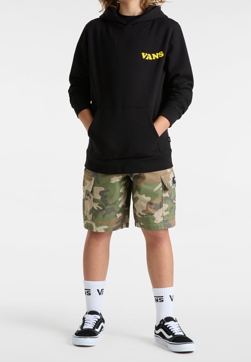 Enfant portant un sweat à capuche Vans noir, un short cargo camouflage, des chaussettes Vans blanches et des baskets Vans noires, debout les mains dans les poches.