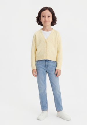 Niño con cabello corto y oscuro que lleva un cárdigan abotonado de color amarillo pálido, camisa blanca, vaqueros azul claro y zapatillas blancas, de pie sobre un fondo blanco.