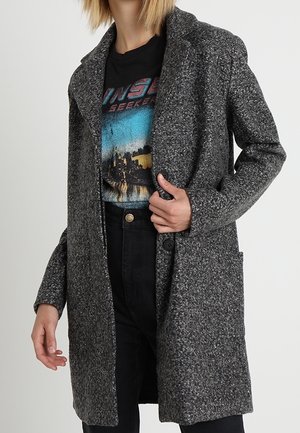 Personne portant un manteau gris texturé sur un t-shirt noir à motif graphique et un pantalon noir, debout devant un fond clair uni.
