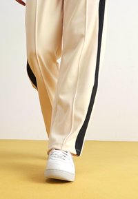 Krämfärgade sportbyxor med svarta sidostråk, gjorda av ett slätt och lätt material. Bärs med vita sneakers.