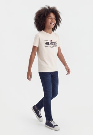 Χαμογελαστό παιδί που περπατά, φορώντας μια μπεζ μπλούζα Hilfiger, σκούρο μπλε τζιν και ναυτικά αθλητικά παπούτσια, με φόντο ένα απλό λευκό φόντο.