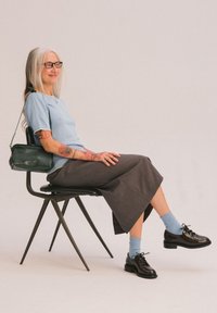 Top celeste a maniche corte, gonna grigia di lunghezza media, calzini blu, scarpe nere con lacci, borsa a tracolla verde, seduta su una sedia nera.