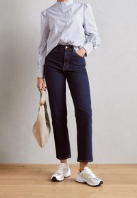 Chemise à rayures bleues et blanches avec des poignets froncés, jeans noirs taille haute et baskets blanches, tenant un sac fourre-tout beige.