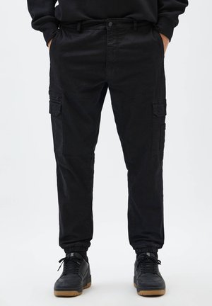 Person trägt schwarze Cargohosen mit Seitentaschen und schwarze Sneakers mit Gummisohlen, steht mit den Händen in den Taschen vor einem schlichten Hintergrund.