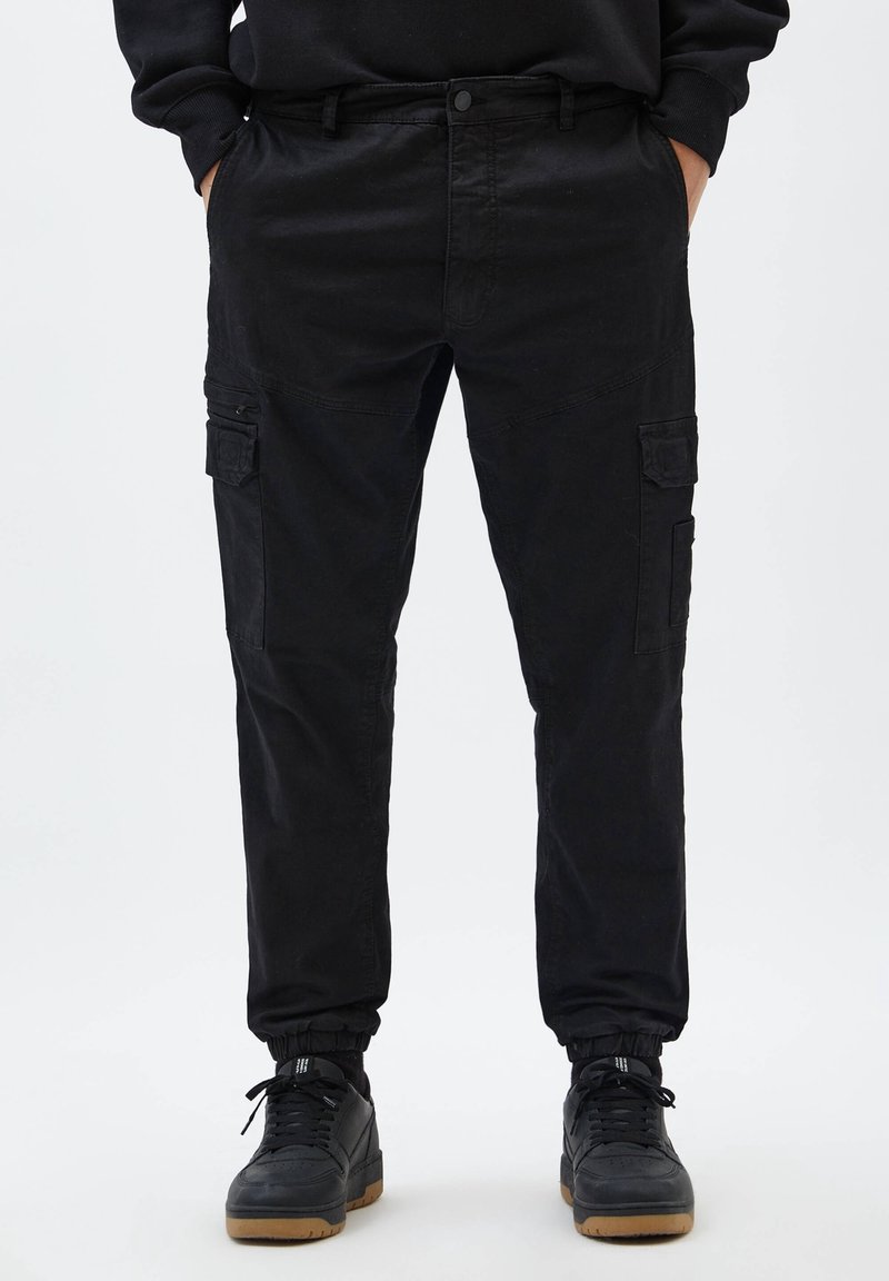 Personne portant un pantalon cargo noir avec des poches latérales et des baskets noires à semelles en gomme, debout les mains dans les poches devant un fond uni.
