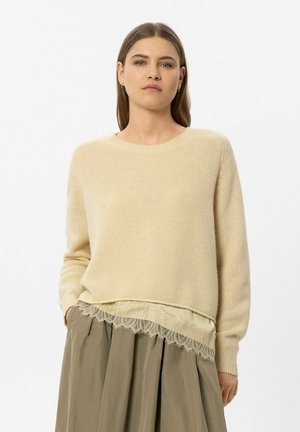 Frau mit glattem braunem Haar trägt einen beigen Strickpullover über einem Spitzen-Top und einem olivgrünen plissierten Rock vor schlichtem Hintergrund.