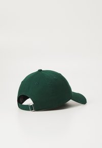 Grønn bomullslue med buet skjerm, justerbar stropp bak og ventilasjonsmuligheter nær toppen. Enkelt design uten logoer.