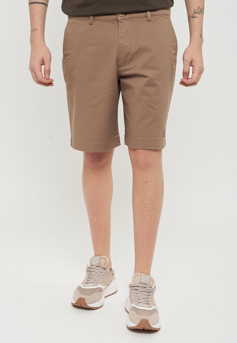 Pantaloni corti in cotone marrone con taglio dritto, tasche laterali e orli arrotolati. Abbinati a sneakers beige. Design minimalista, senza fantasie.