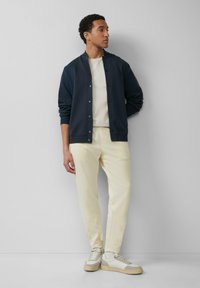 Marine sweatjack met knopen, crèmekleurig t-shirt, lichtbeige broek en witte sneakers met grijze accenten, tegen een neutrale achtergrond.