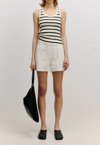 Gestreiftes schwarzes und weißes Rippen-Top, weiße Shorts, schwarze flache Schlupfschuhe. Hält eine große schwarze Tragetasche. Glatte Texturen überall.