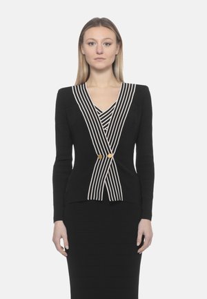 Donna che indossa un blazer nero aderente con righe verticali bianche e bottoni dorati, abbinato a una gonna nera coordinata, in piedi immobile.