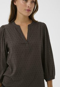 Bruine, gestructureerde blouse met een placket-halslijn en pofmouwen. Het materiaal heeft subtiele verticale strepen en kleine stippenpatronen.