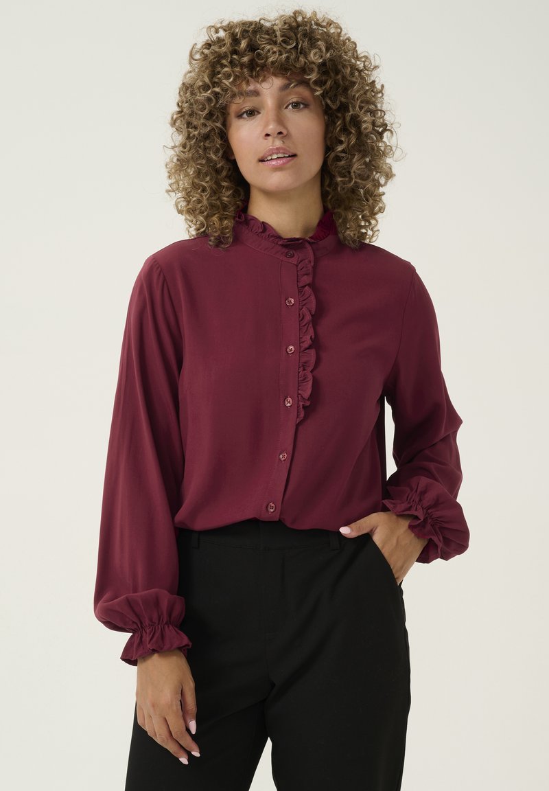 Maroon blouse with a ruffled collar and button front, featuring long sleeves with elastic cuffs. Paired with black trousers.