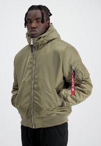 Giacca bomber verde oliva con cappuccio, chiusura con zip, polsini a costine, due tasche frontali e un'etichetta rossa sulla manica sinistra.