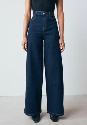 Džíny s vysokým pasem, širokými nohavicemi z tmavého denimu, viditelným švem, zapínáním na knoflík a hladkým povrchem. Mají čistý, minimalistický design.