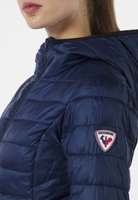 Giubbotto imbottito blu navy con una texture lucida, design a cuciture orizzontali e un patch con il logo Rossignol sulla spalla sinistra. Chiusura frontale con zip.