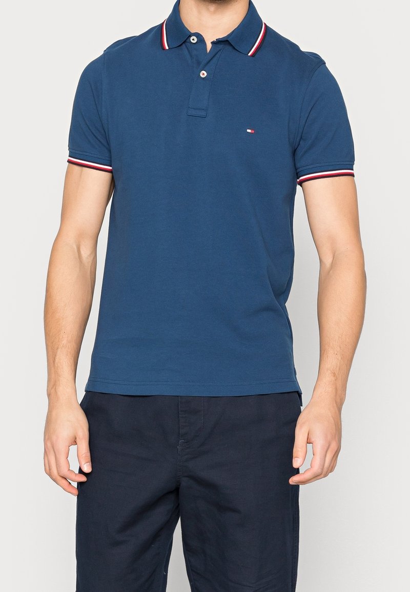 Homme portant un polo bleu à manches courtes avec un col et des manches rayés rouge, blanc et noir, associé à un pantalon bleu marine foncé.
