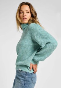 Maglione a collo alto ribbed blu chiaro con maniche ampie e orlo leggermente cropped. Indossato con jeans blu chiaro. Presenta una piccola etichetta sul lato.