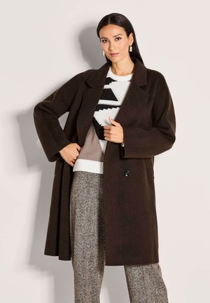 Vrouw in een donkerbruine lange jas over een gebreid patroontrui en grijze herringbone broek, staande tegen een lichte achtergrond.