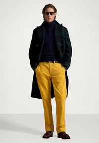 Άνδρας φορώντας σκοτεινό καρό παλτό, μπλε ύφασμα ζιβάγκο, κίτρινες παντελόνες από καστόρι, καφέ loafers και γυαλιά ηλίου, στέκεται με τα χέρια στις τσέπες.