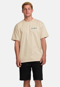 Beige cotton t-shirt with short sleeves, featuring a green logo on the left chest. Worn with black shorts, displayed on a neutral background.