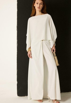Femme portant une combinaison blanche à jambes larges avec un haut drapé, tenant un sac à main beige tissé, debout devant un fond noir et blanc.