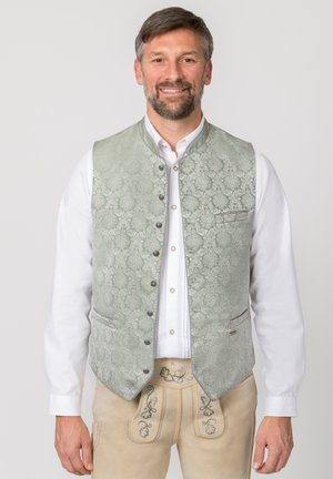 Lächelnder Mann, trägt ein weißes Hemd mit Knöpfen, eine gemusterte hellgrüne Weste und beige traditionelle bestickte Hosen.