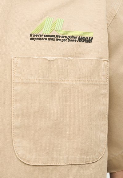 Μπεζ ύφασμα με ραμμένη τσέπη και πράσινη και μαύρη κεντητή γραφή που λέει "Φαίνεται πως ποτέ δεν πηγαίνουμε πουθενά μέχρι να φτάσουμε εκεί MSGM."