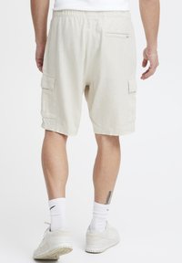 Beige cargo shorts met een elastische tailleband, zijzakken en een achterzak met rits. Draagt met witte sneakers en sokken.