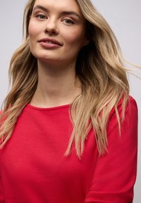 Jonge vrouw met lang blond haar, gekleed in een rood shirt, kijkt licht omhoog met een zachte glimlach tegen een effen lichte achtergrond.