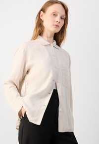 Beige, pitkähihainen paita, jossa on kauluskangas, etuvetskaus ja rintatasku; valmistettu kevyestä, teksturoidusta kankaasta.