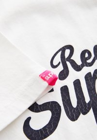 Närbild av vitt tyg med svart strukturerat tryck som säger "Super" och en liten rosa tygetikett på sömmen.