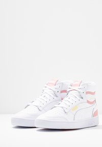 Bílé vysoké tenisky s růžovými a stříbrnými akcenty. Mezi rysy patří texturovaný kožený svršek, perforovaná špička a gumová podrážka s vzorovaným profilováním.