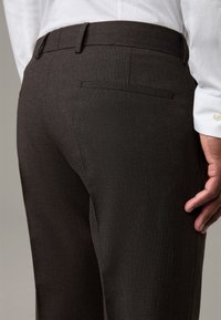 Dunkelbraune maßgeschneiderte Hosen aus strukturiertem Stoff, mit einer Gesäßtasche und einem glatten Bund. Kombiniert mit einem weißen Hemd.
