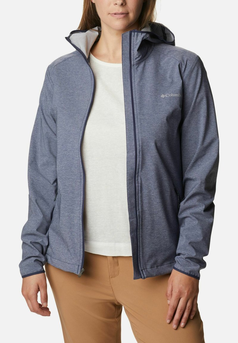 Giacca leggera blu Columbia con cappuccio, chiusura con zip e polsini regolabili, realizzata in un tessuto morbido. Indossata sopra una camicia leggera e pantaloni.