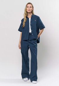 Mörkblå kortärmad skjorta med knappar över en vit linne, kombinerad med matchande vida byxor. Modellen bär vita skor.