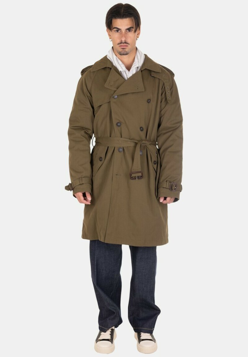 Trench coat verde oliva con design doppiopetto, grandi bottoni, spalline e cintura legata; abbinato a jeans scuri e sneakers.