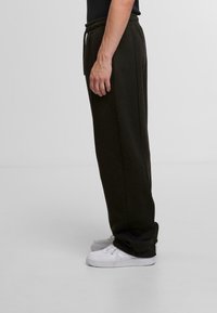 Dark green sweatpants with an elastic waist and side pockets, displayed on a person wearing white sneakers, viewed from the side.