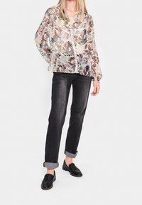 Genomskinlig blommig blus i pastellfärger med långa ärmar och knäppning, parat med mörka jeans och svarta slip-on skor med en strukturerad design.