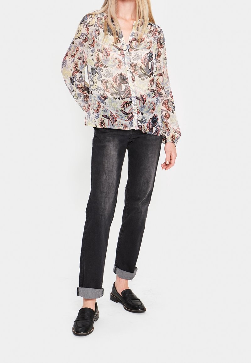 Genomskinlig blommig blus i pastellfärger med långa ärmar och knäppning, parat med mörka jeans och svarta slip-on skor med en strukturerad design.