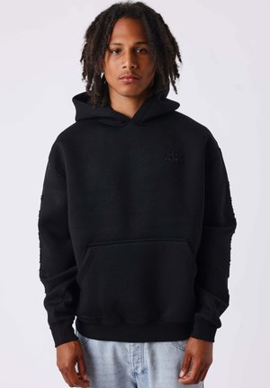Jeune homme aux cheveux bouclés mi-longs, portant un sweat à capuche noir et un jean bleu clair, debout devant un fond blanc uni.