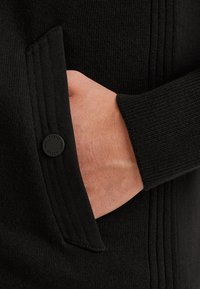 Mustat neuleet, joissa on uritettu pinta ja sivutaskut. Lähellä taskua on painike, jossa on "PINE LEGEND" -brändäys. Pehmeä neulottu materiaali.