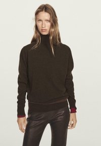 Frau mit langen blonden Haaren, die einen schwarzen Rollkragenpullover mit rotgestreiften Bündchen und schwarze Lederhosen vor einem einfarbigen Hintergrund trägt.