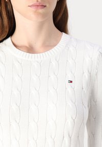 Biały sweter w splot kabelkowy z okrągłym dekoltem, z małym haftowanym logo w czerwonym, białym i niebieskim kolorze na piersi.