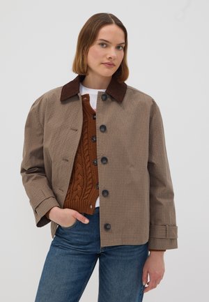 Woman wearing a brown checkered jacket with large buttons, a rust cable-knit vest, white shirt, and blue jeans, posing with one hand in pocket.