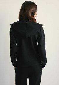 Kobieta z brązowymi włosami, ubrana w czarną bluzę z kapturem i czarne spodnie, stoi przodem do jasnej ściany, z rękami w kieszeniach.