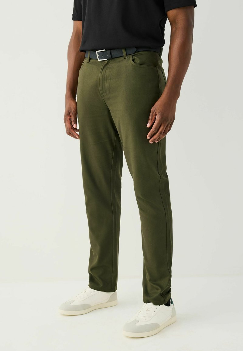 Olivově zelené, úzké kalhoty z hladké látky mají přední kapsy, poutka na opasek a jsou stylizované s černým pásem a bílými teniskami.