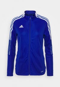 Veste de sport Adidas bleue zippée avec des bandes blanches sur les manches et le logo Adidas blanc sur la poitrine, présentée sur un fond uni.