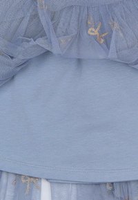 Top azzurro chiaro fatto di un tessuto morbido, stratificato con tulle trasparente con motivi di fiocchi dorati lungo l'orlo. Finitura liscia con dettagli arricciati.