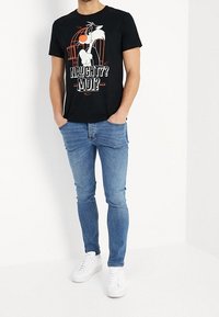 Hombre con una camiseta negra que tiene un personaje de dibujos animados y el texto "¿Travieso? ¿Yo?" combinada con unos jeans ajustados azules y zapatillas blancas, de pie con las manos en los bolsillos.